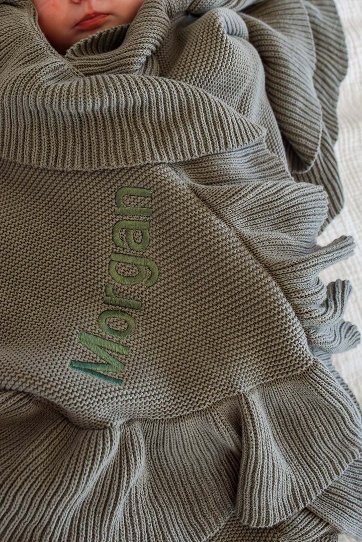 A close-up of a baby swaddled in a ribbed knit blanket with ruffled edges and the name 'Morgan' embroidered vertically in olive green.