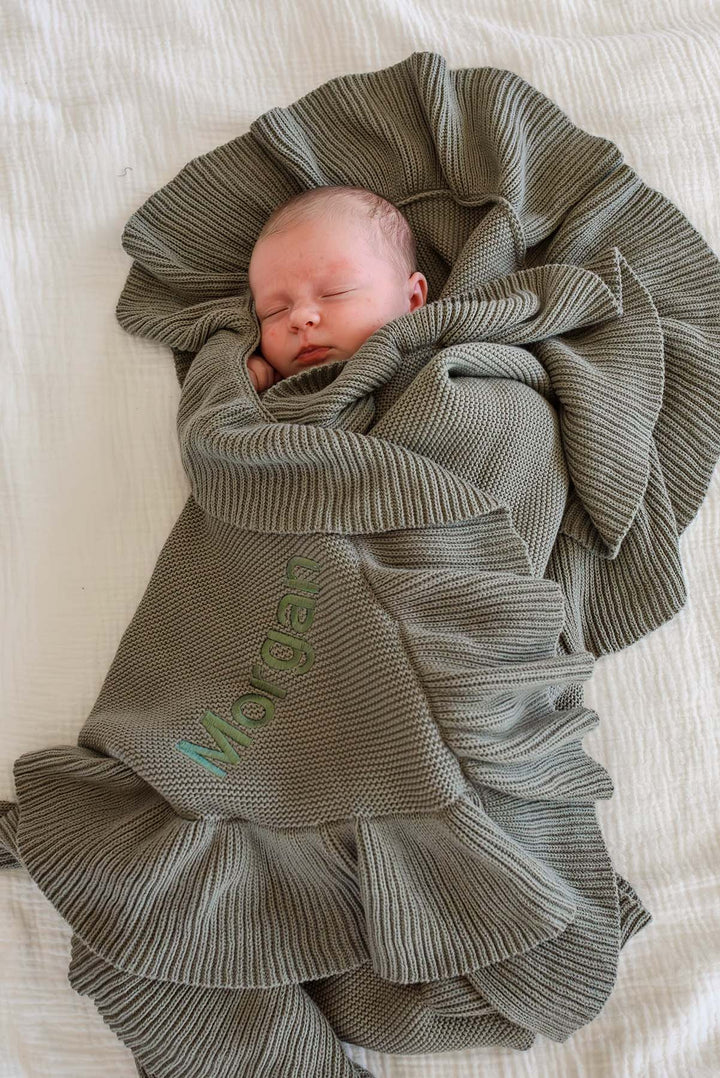A sleeping newborn baby swaddled in a textured olive green knit blanket with ruffled edges, lying on a white quilted bed. The name 'Morgan' is embroidered in a light green thread on the blanket.