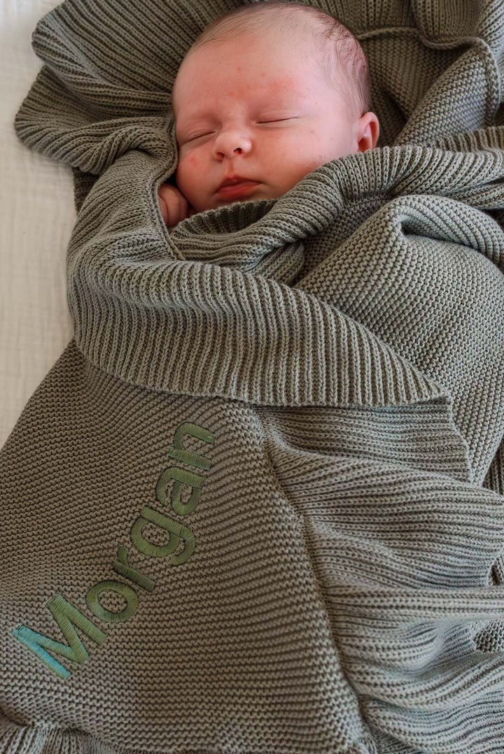 A sleeping newborn baby wrapped in a textured olive green knit blanket with the name 'Morgan' embroidered in pastel green and blue on the front.