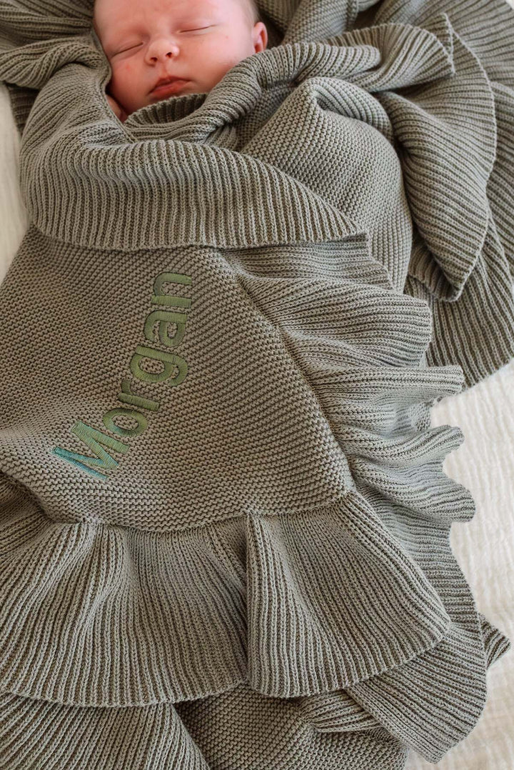A close-up of a sleeping newborn baby wrapped in a textured olive green knit blanket with ruffled edges and personalized embroidery reading 'Morgan'.