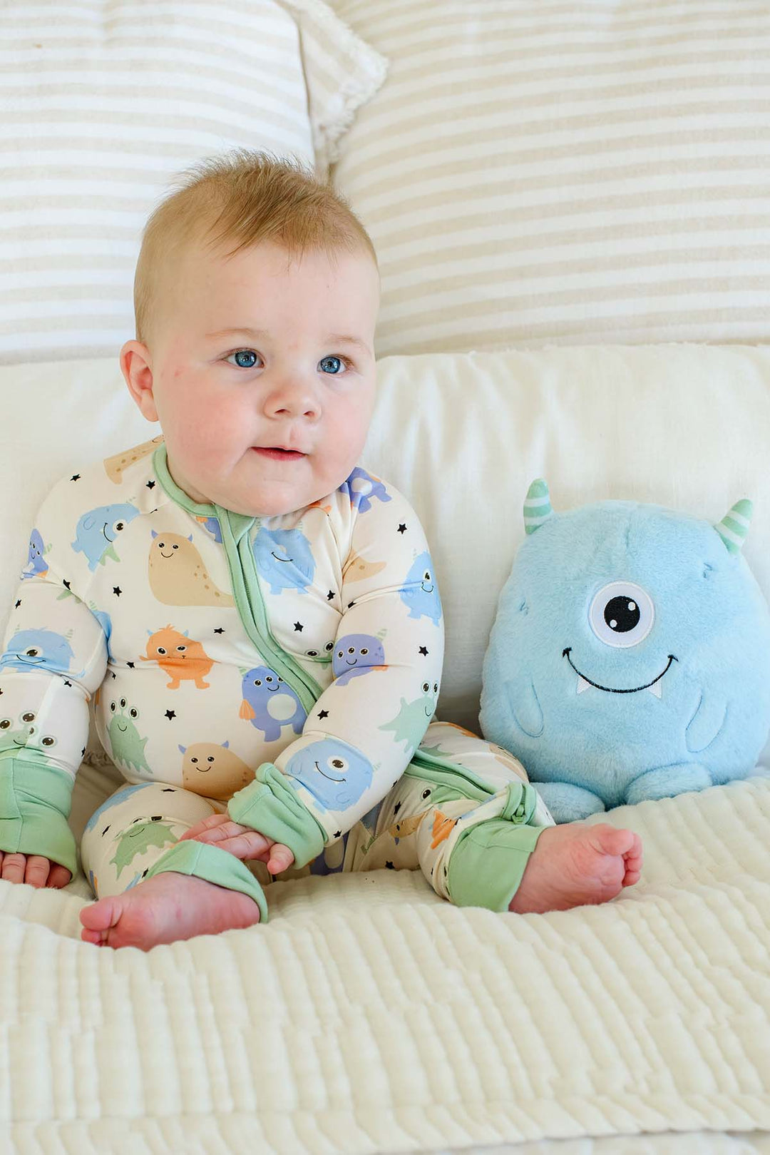 Baby sitting on a bed next to a plush monster toy, wearing a Caden Lane Little Monsters zipper footie with sage green trim, showing blue, green, and orange cartoon monsters