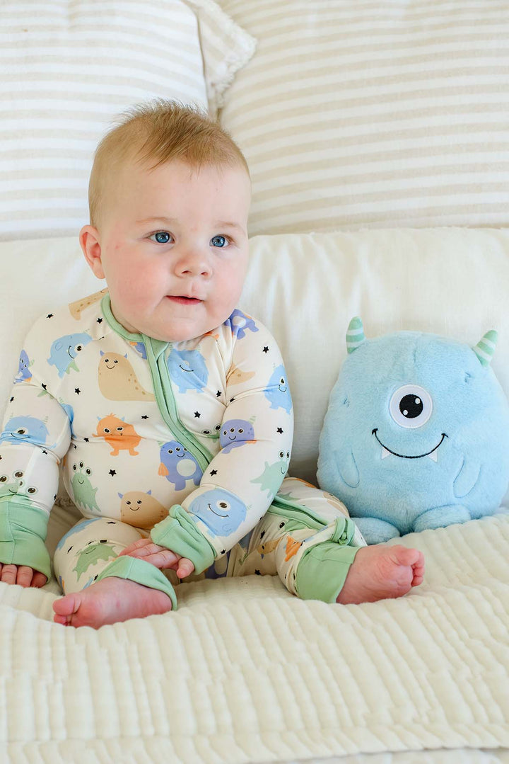 Baby sitting on a bed next to a plush monster toy, wearing a Caden Lane Little Monsters zipper footie with sage green trim, showing blue, green, and orange cartoon monsters