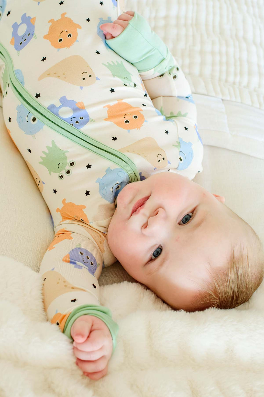 Close-up of a baby lying down smiling, wearing a Caden Lane Little Monsters zipper footie, showing print detail and sage green trim