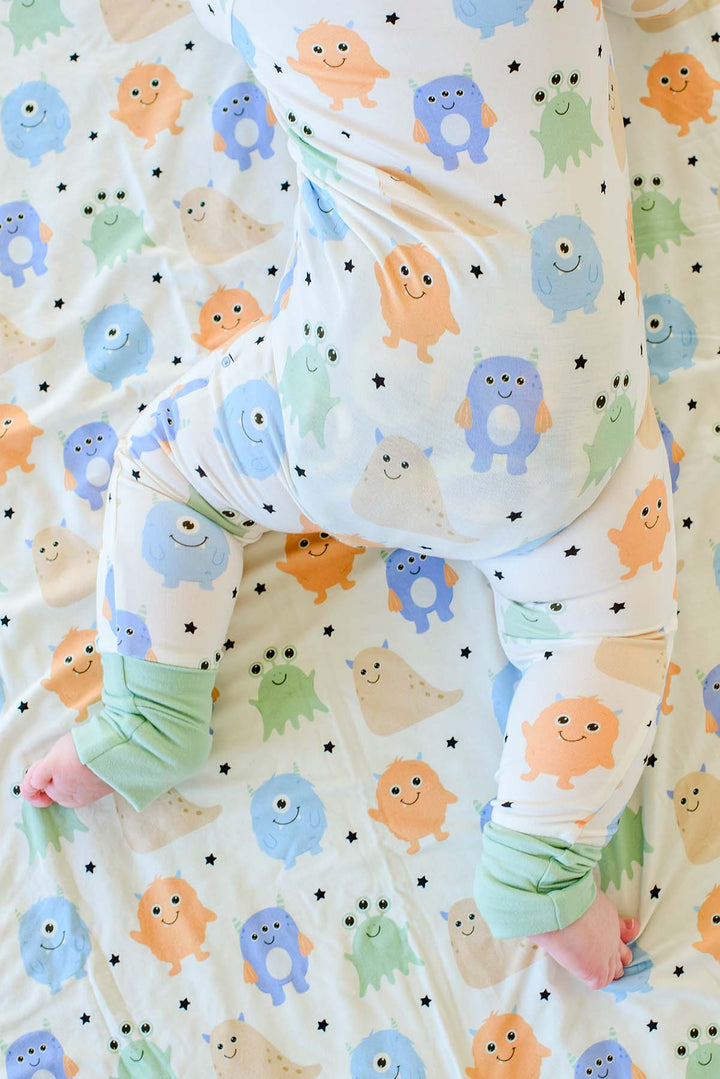 Close-up of baby crawling in Caden Lane Little Monsters print pajamas with blue, orange, tan, and green cartoon monsters and stars, sage green cuffs