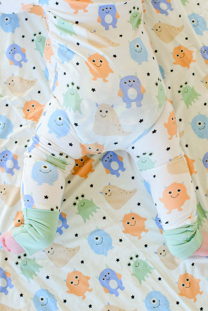 Close-up of a baby's legs and feet from above in a Caden Lane Little Monsters zipper footie, showing the print and sage green footed cuffs