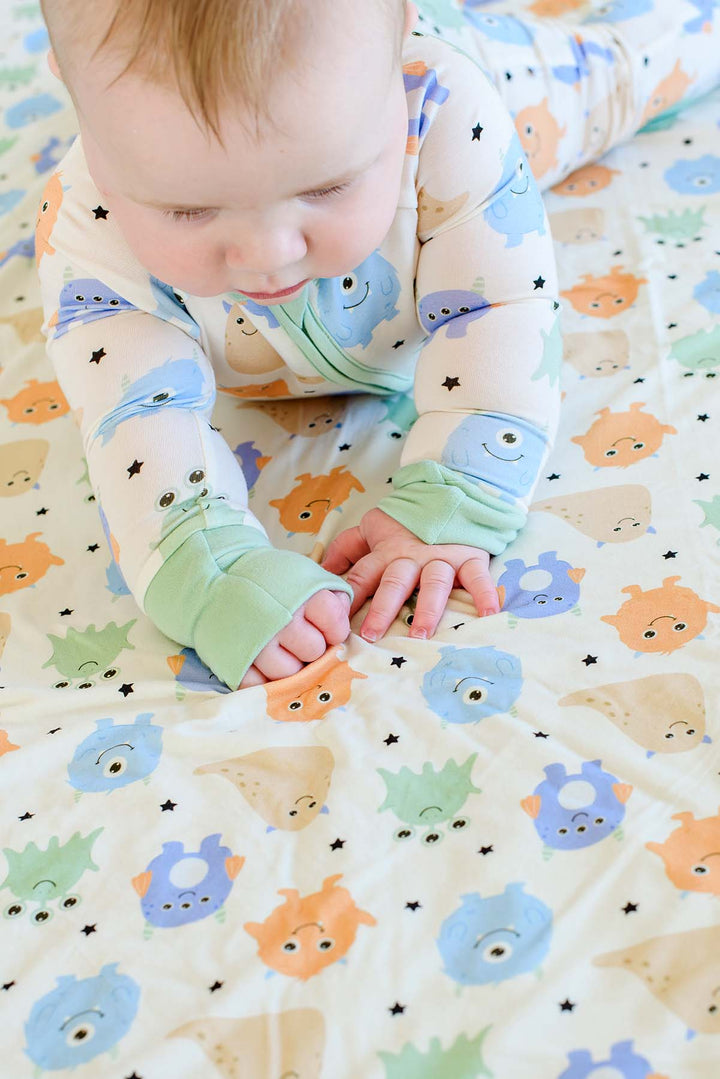 Baby crawling on coordinating monster-print bedding, wearing a Caden Lane Little Monsters zipper footie with sage green trim