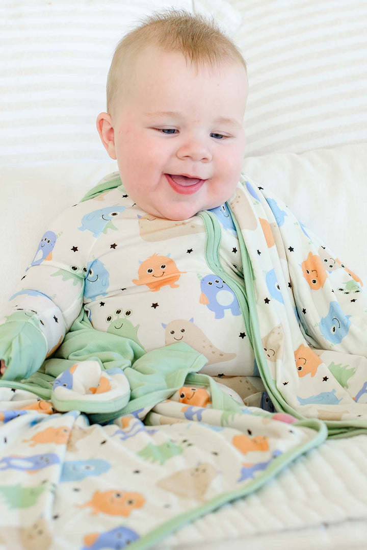 Smiling baby wrapped in a Caden Lane sage green-trimmed Little Monsters bamboo blanket, wearing matching monster-print pajamas