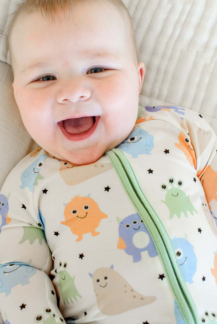 Close-up of a laughing baby lying down, wearing a Caden Lane Little Monsters zipper footie, showing print detail and sage green zipper trim