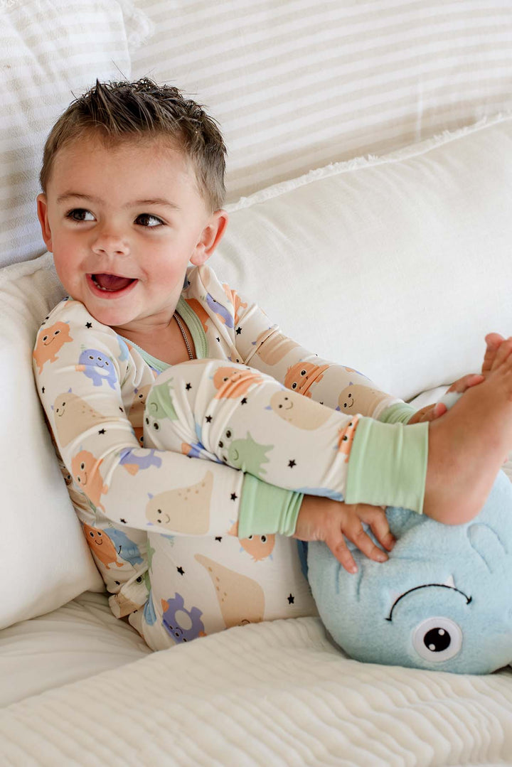 Smiling toddler curled up on a couch with a plush monster toy, wearing a Caden Lane Little Monsters two-piece pajama set with sage green trim