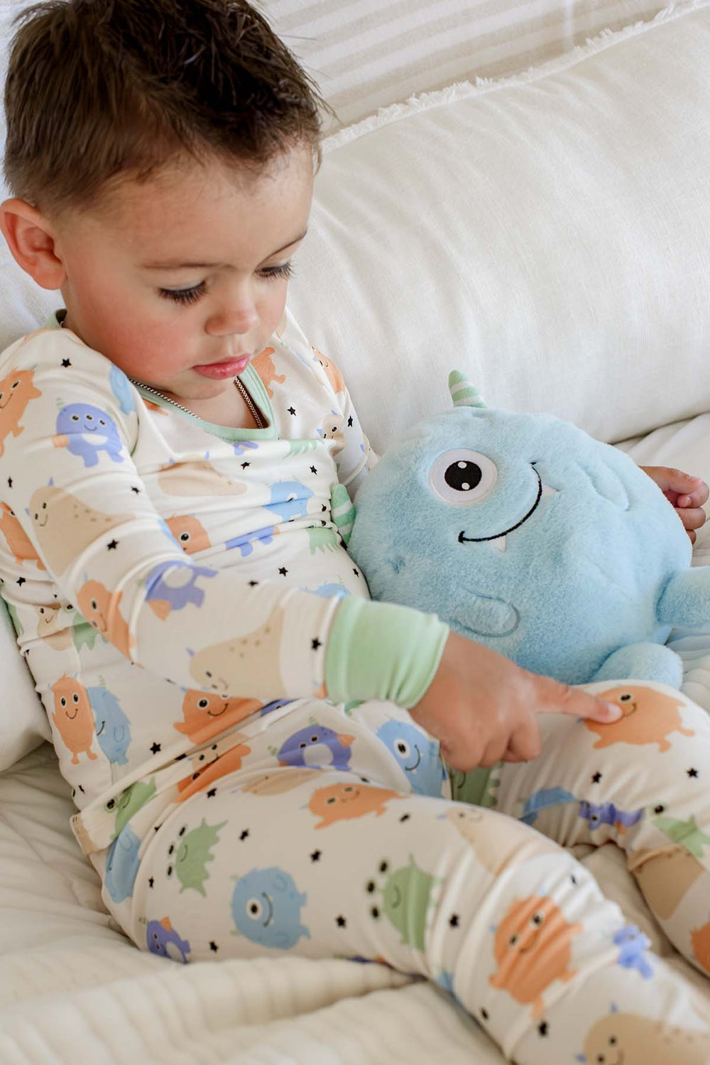 Toddler sitting on a couch pointing at a plush monster toy, wearing a Caden Lane Little Monsters two-piece pajama set, showing print detail and sage green cuffs