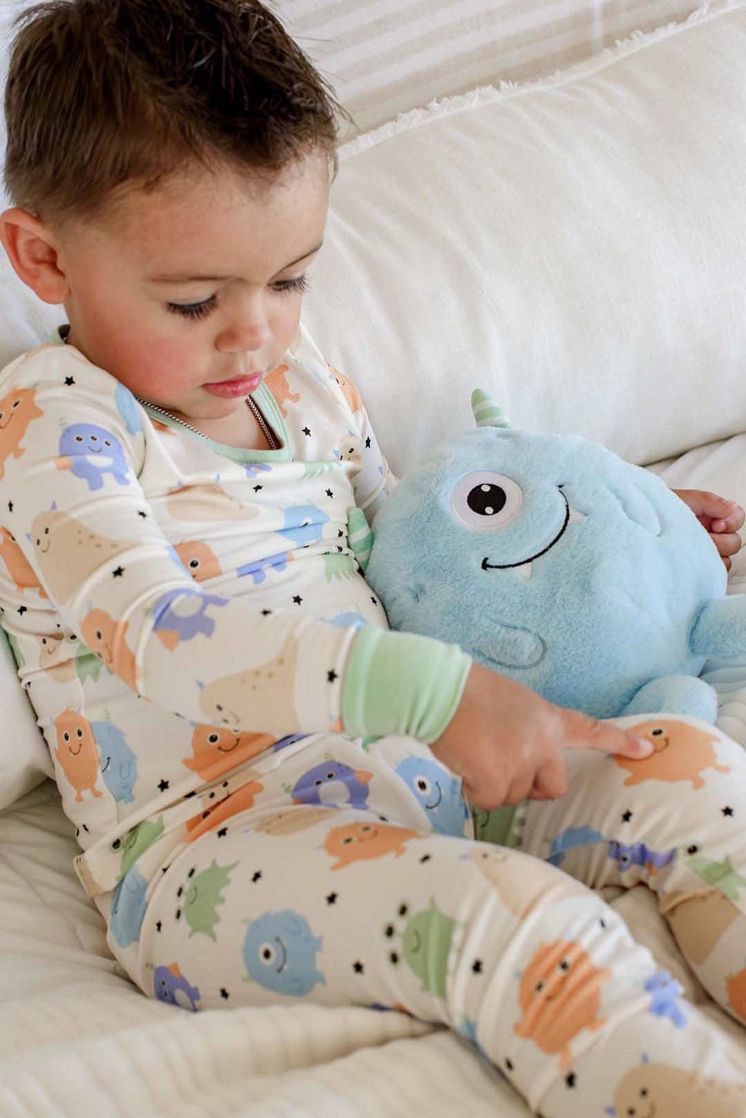 Toddler sitting on a couch pointing at a plush monster toy, wearing a Caden Lane Little Monsters two-piece pajama set, showing print detail and sage green cuffs