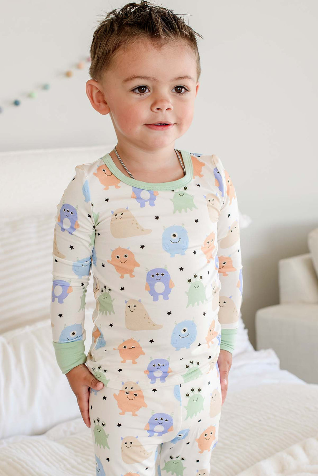 Toddler standing on a bed wearing a Caden Lane Little Monsters two-piece pajama set with sage green trim, full front view
