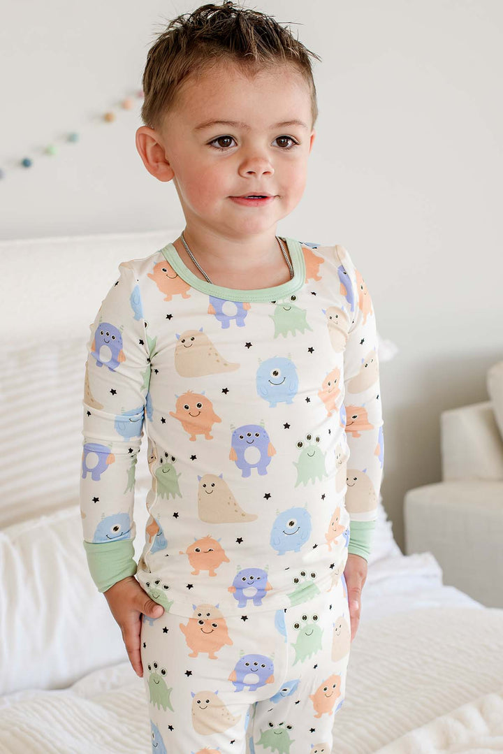 Toddler standing on a bed wearing a Caden Lane Little Monsters two-piece pajama set with sage green trim, full front view