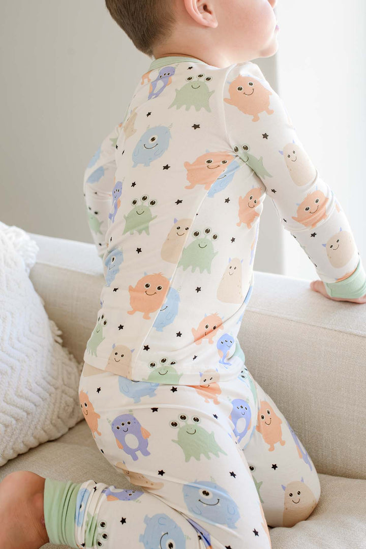 Toddler kneeling on a couch, back view showing print detail and sage green cuffs of a Caden Lane Little Monsters two-piece pajama set