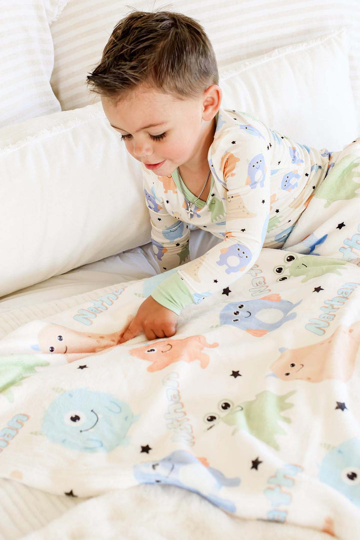 Toddler boy in matching monster-print pajamas playing with a Caden Lane personalized "Nathan" Little Monsters minky blanket on a bed