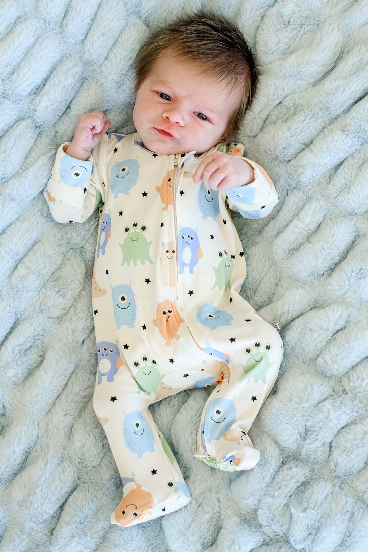 Newborn laying on furry blue blanket wearing a Caden Lane Little Monsters zipper footie in cream with blue, green, and orange cartoon monsters