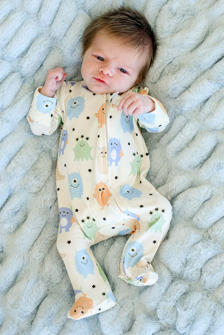 Newborn laying on furry blue blanket wearing a Caden Lane Little Monsters zipper footie in cream with blue, green, and orange cartoon monsters
