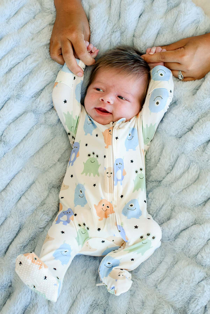 Smiling newborn held by parents' hands, wearing a Caden Lane Little Monsters zipper footie