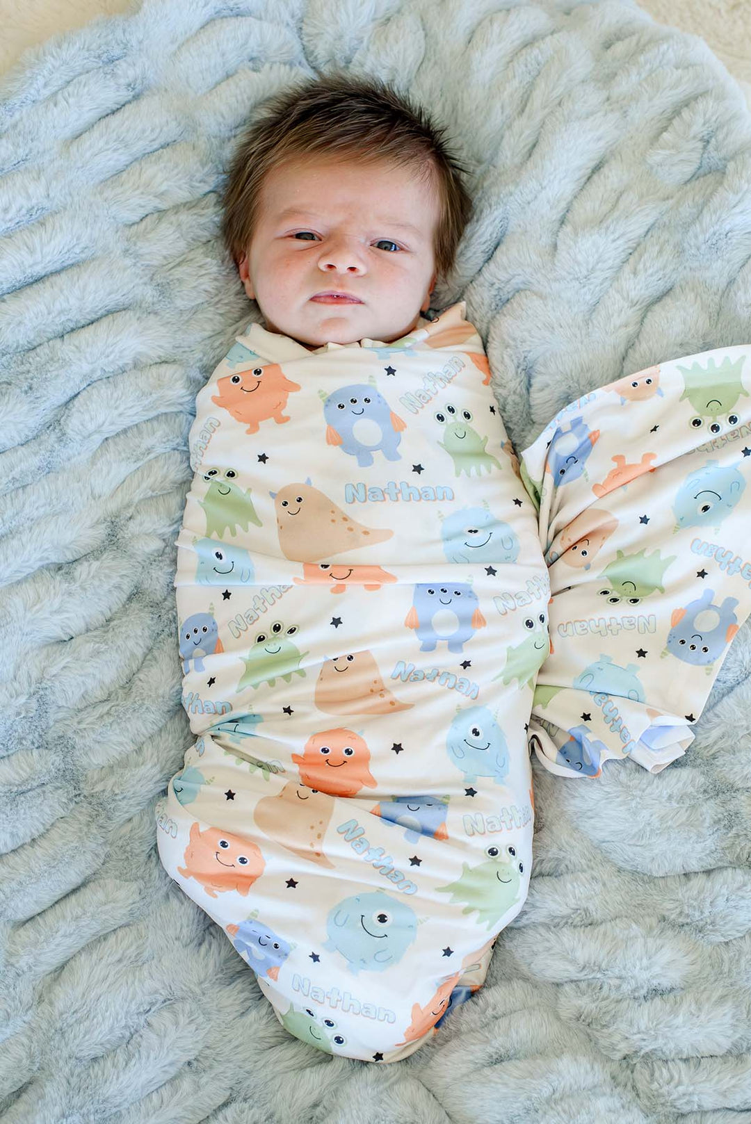 Newborn swaddled in Caden Lane personalized "Nathan" Little Monsters print blanket, close-up view on a blue faux fur blanket