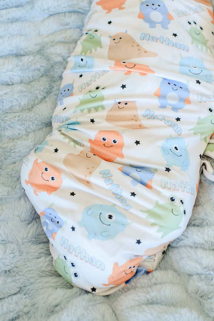Close-up of Caden Lane personalized "Nathan" swaddle blanket in Little Monsters print with blue, green, orange, and tan cartoon monsters and stars on a blue faux fur blanket