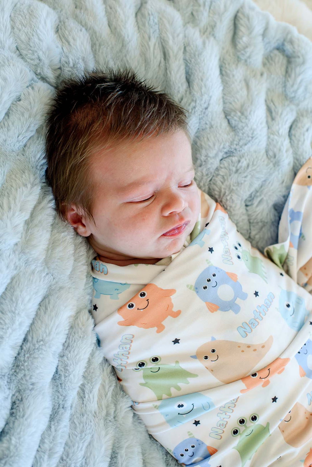 Sleeping newborn swaddled in Caden Lane personalized "Nathan" Little Monsters print blanket with blue, green, orange, and tan cartoon monsters, laying on a blue faux fur blanket