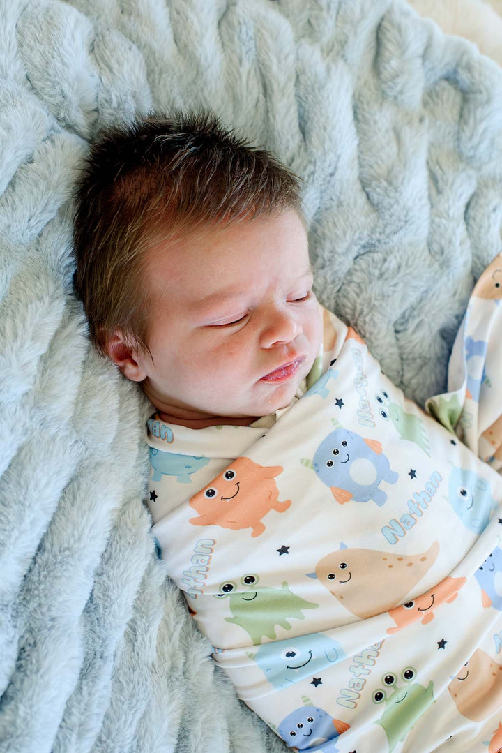 Sleeping newborn swaddled in Caden Lane personalized "Nathan" Little Monsters print blanket with blue, green, orange, and tan cartoon monsters, laying on a blue faux fur blanket