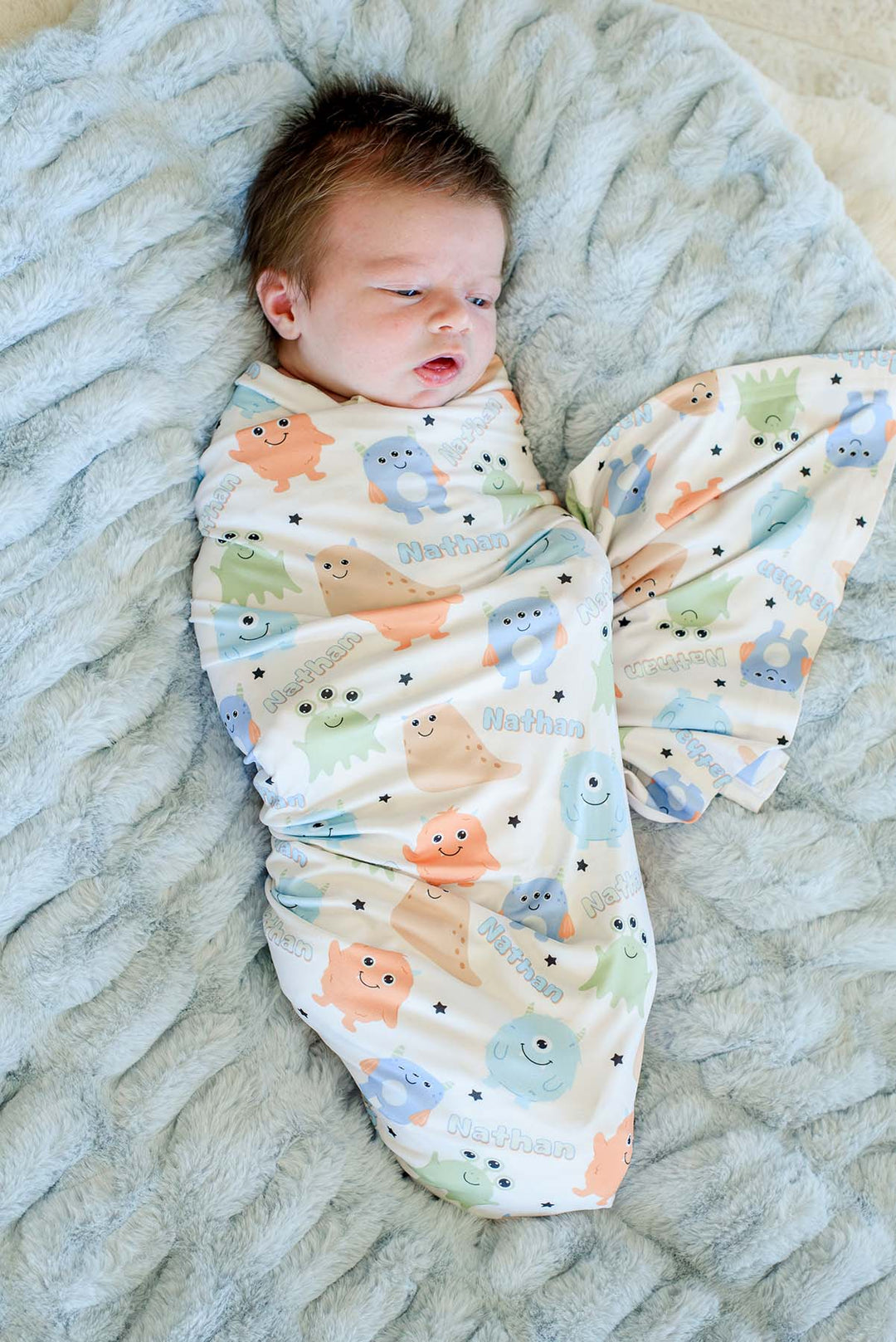 Newborn swaddled in Caden Lane personalized "Nathan" Little Monsters print blanket, overhead view on a blue faux fur backdrop