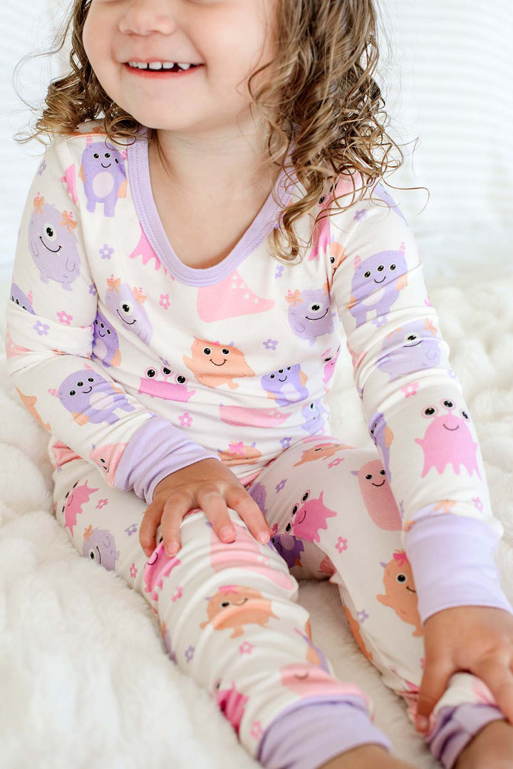 Close-up of a toddler sitting cross-legged, wearing a Caden Lane Little Monsters two-piece pajama set with lavender trim, showing pink, purple, and orange cartoon monsters on cream fabric