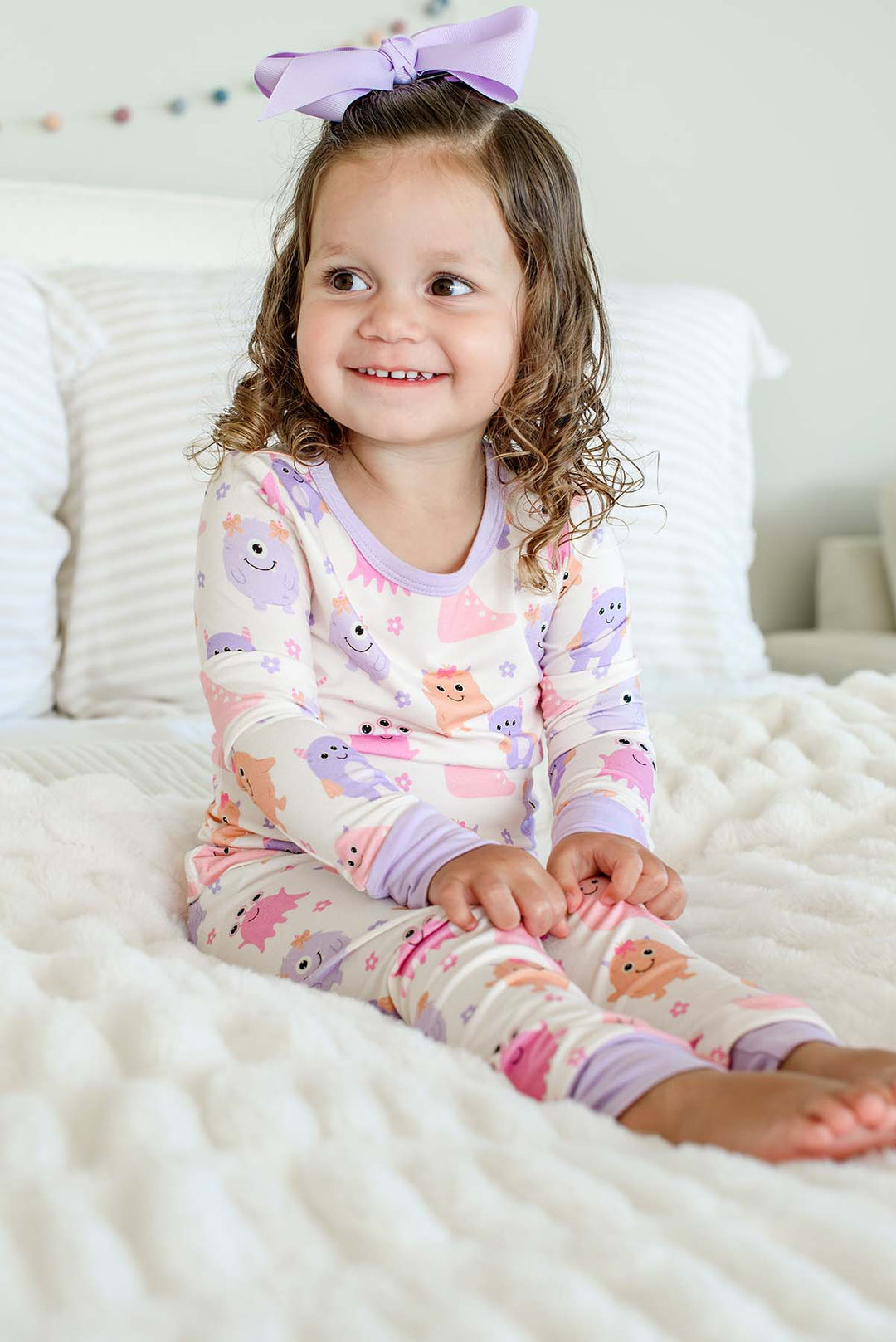 Smiling toddler sitting on a bed wearing a Caden Lane Little Monsters two-piece pajama set with a matching lavender bow, full front view