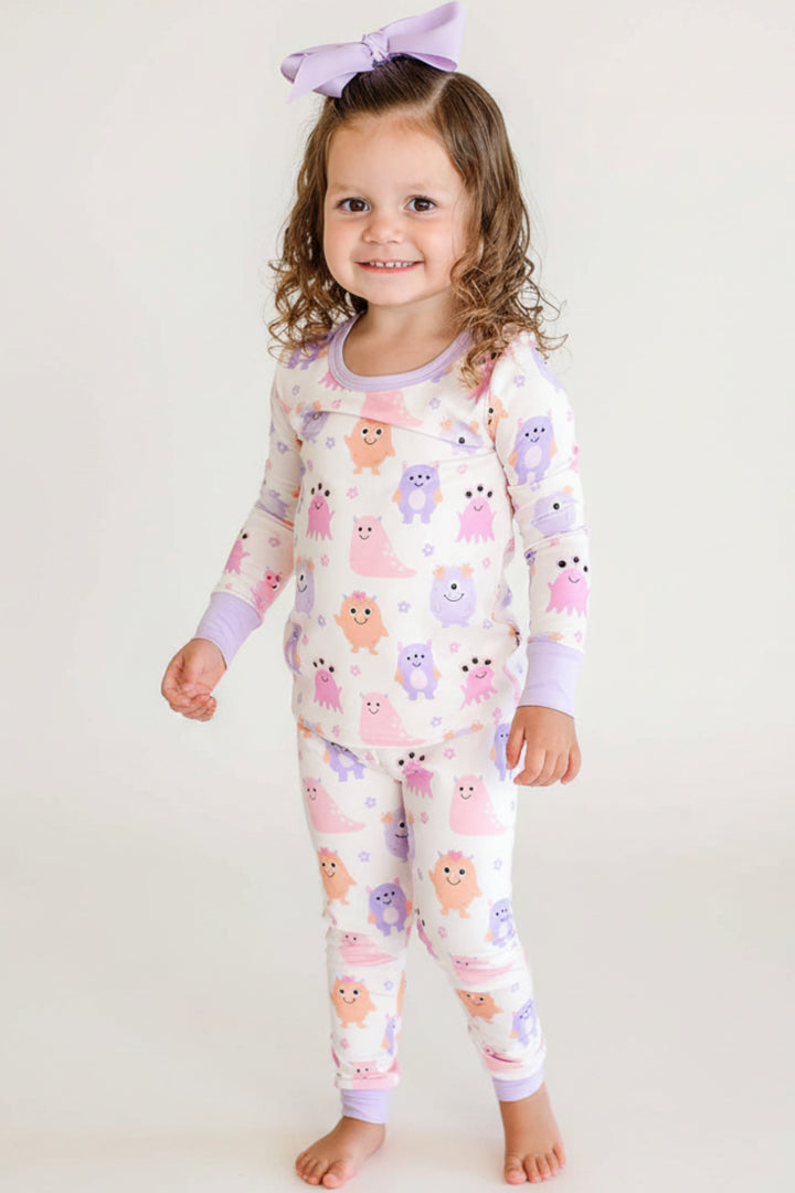 Toddler standing on a bed wearing a Caden Lane Little Monsters two-piece pajama set with matching lavender bow, full front view