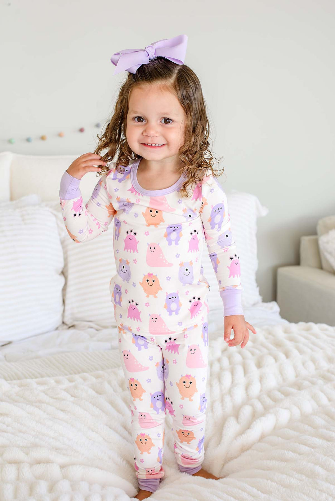 Toddler standing on a bed wearing a Caden Lane Little Monsters two-piece pajama set with matching lavender bow, full front view