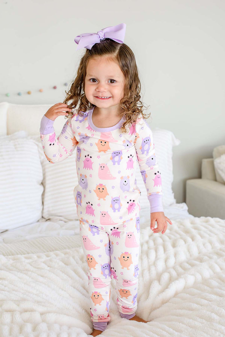 Toddler standing on a bed wearing a Caden Lane Little Monsters two-piece pajama set with matching lavender bow, full front view