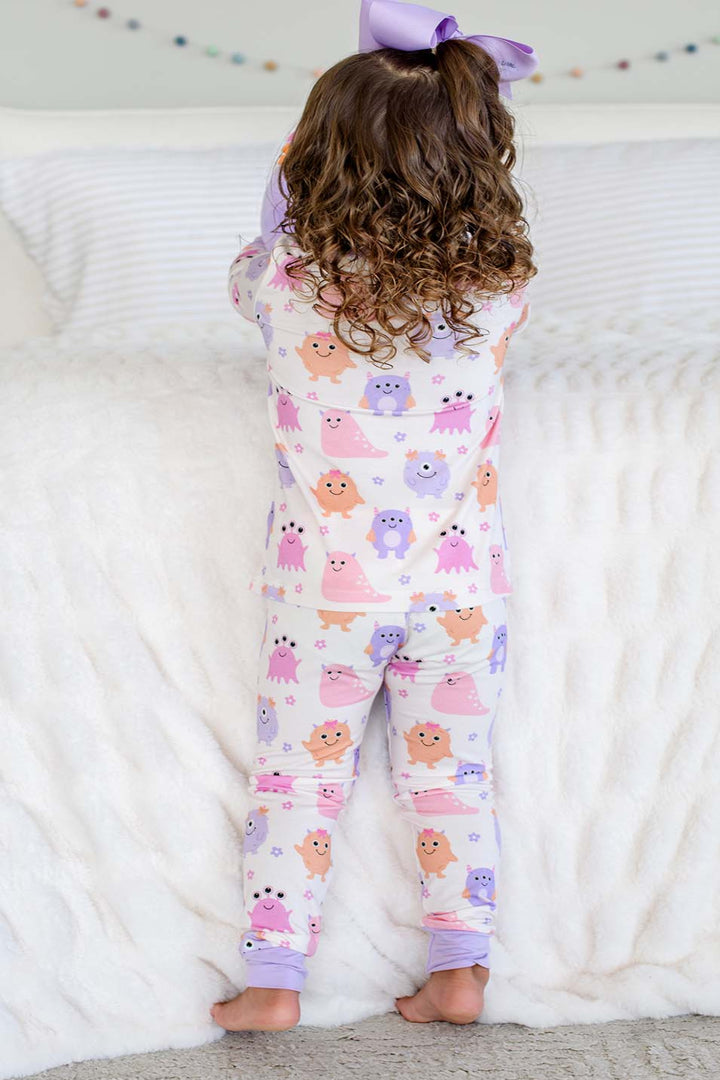 Toddler standing on a bed wearing a Caden Lane Little Monsters two-piece pajama set with matching lavender bow, back view showing print detail