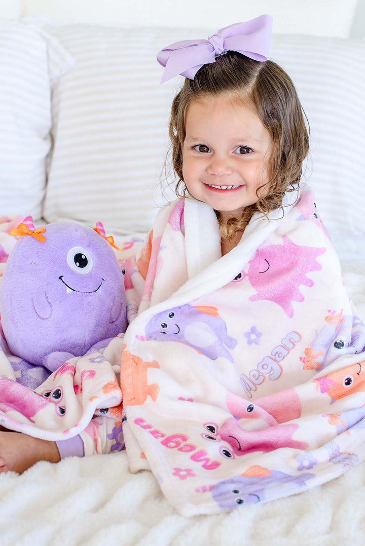 Toddler girl in a lavender bow wrapped in a Caden Lane personalized "Megan" Little Monsters minky blanket, holding a matching purple monster plush