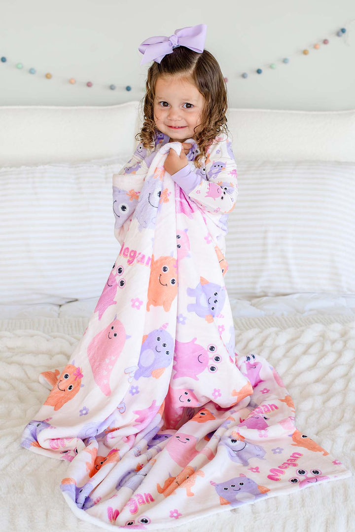 Toddler girl in a lavender bow standing on a bed wrapped in a Caden Lane personalized "Megan" Little Monsters minky blanket