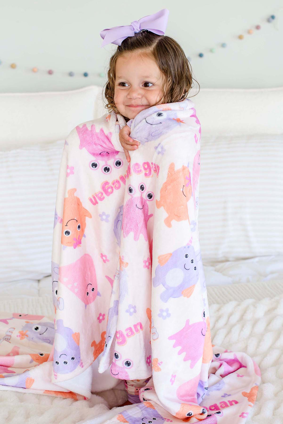 Toddler girl in a lavender bow standing on a bed wrapped in a Caden Lane personalized "Megan" Little Monsters minky blanket