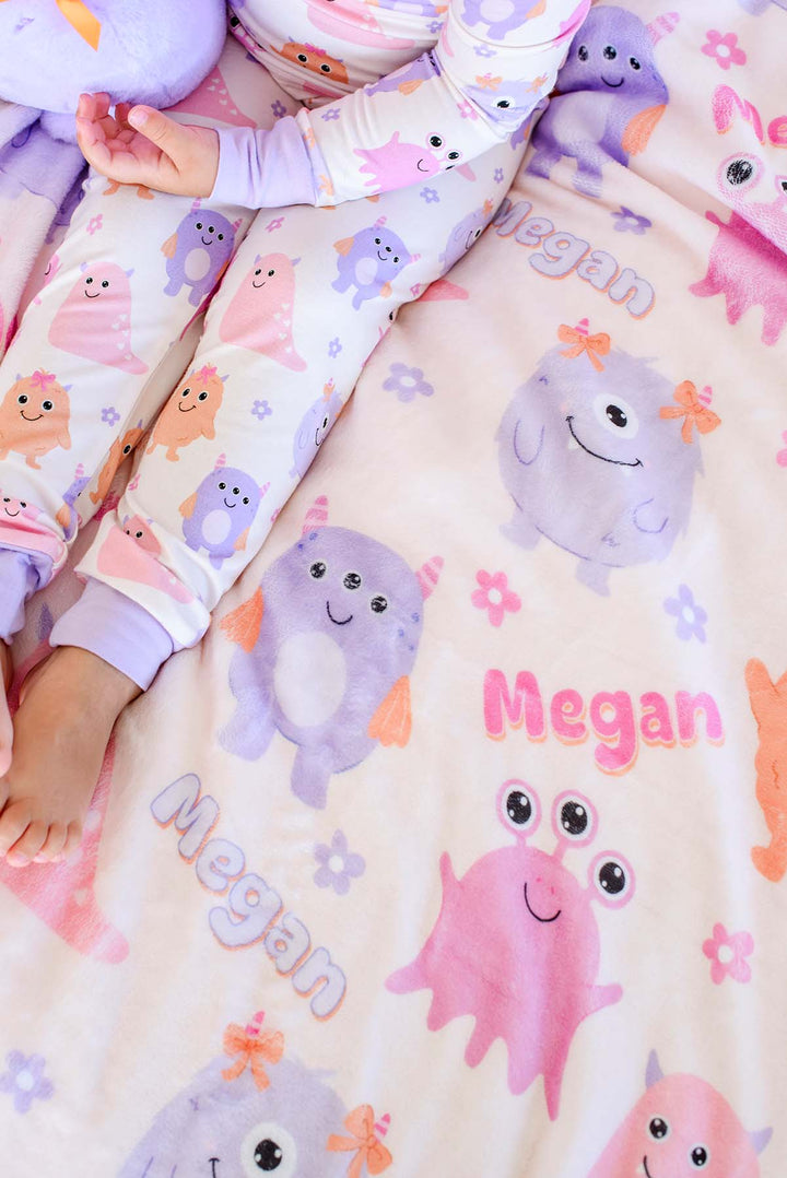 Close-up of a toddler's legs beside a Caden Lane personalized "Megan" Little Monsters minky blanket with pink, purple, and orange cartoon monsters and flowers