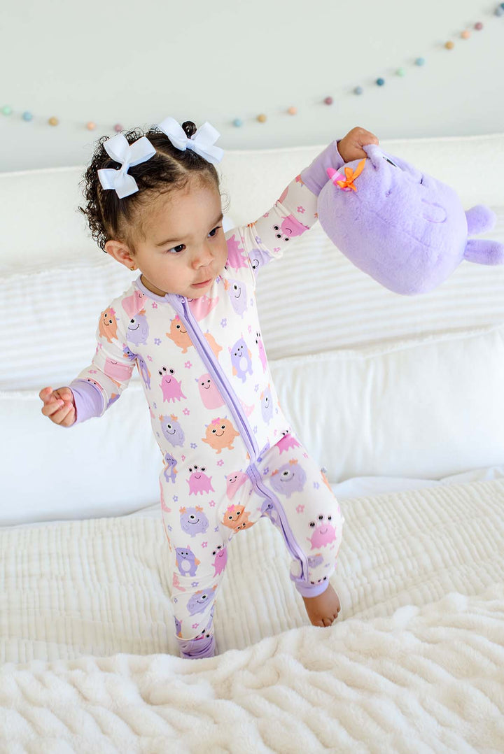 Toddler standing on a bed holding a plush monster toy, wearing a Caden Lane Little Monsters zipper footie with lavender trim, showing pink, purple, and orange cartoon monsters