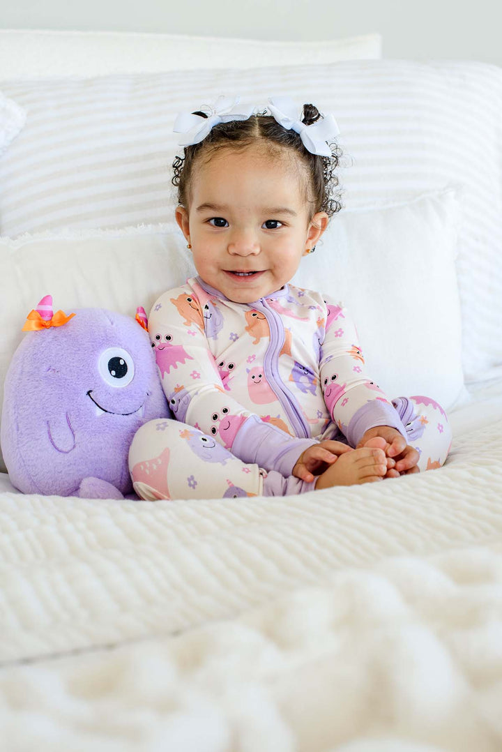 Smiling toddler sitting on a bed with a plush monster toy, wearing a Caden Lane Little Monsters zipper footie with matching bow accents