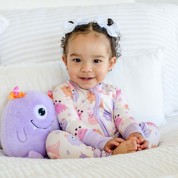Smiling toddler in white hair bows, wearing a Caden Lane pink bamboo zipper romper in a Mini Monsters Pink print featuring friendly monsters, paired with a purple monster plush.