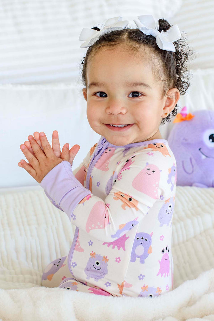 Smiling toddler sitting on a bed clapping, wearing a Caden Lane Little Monsters zipper footie, with a plush monster toy in the background