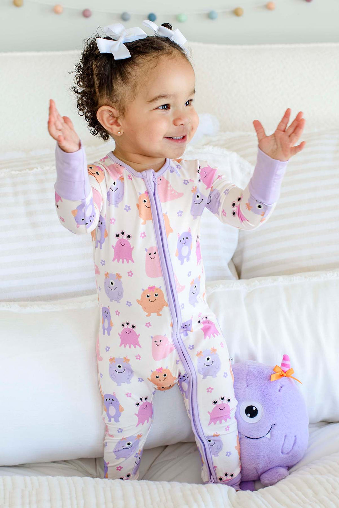 Toddler standing on a bed with a plush monster toy, wearing a Caden Lane Little Monsters zipper footie, full front view