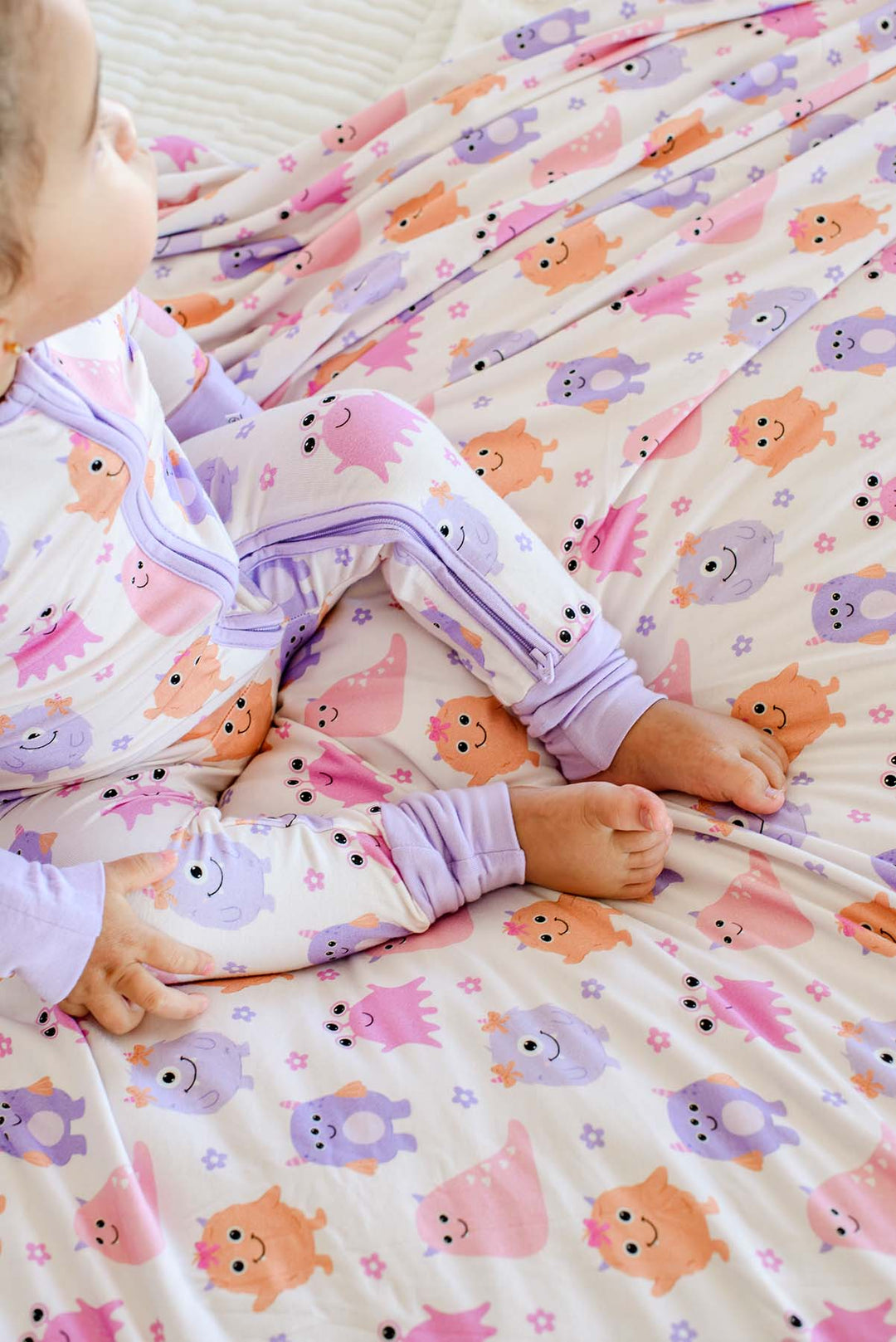 Close-up of a baby's legs in Caden Lane Little Monsters print pajamas with lavender zipper trim, laying on a matching print blanket