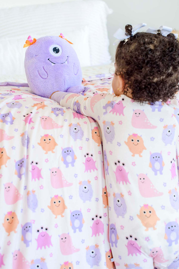 Little girl reaching for the cute Caden Lane monster sitting on a bed, wearing a Caden Lane Little Monsters zipper footie with matching bow accents