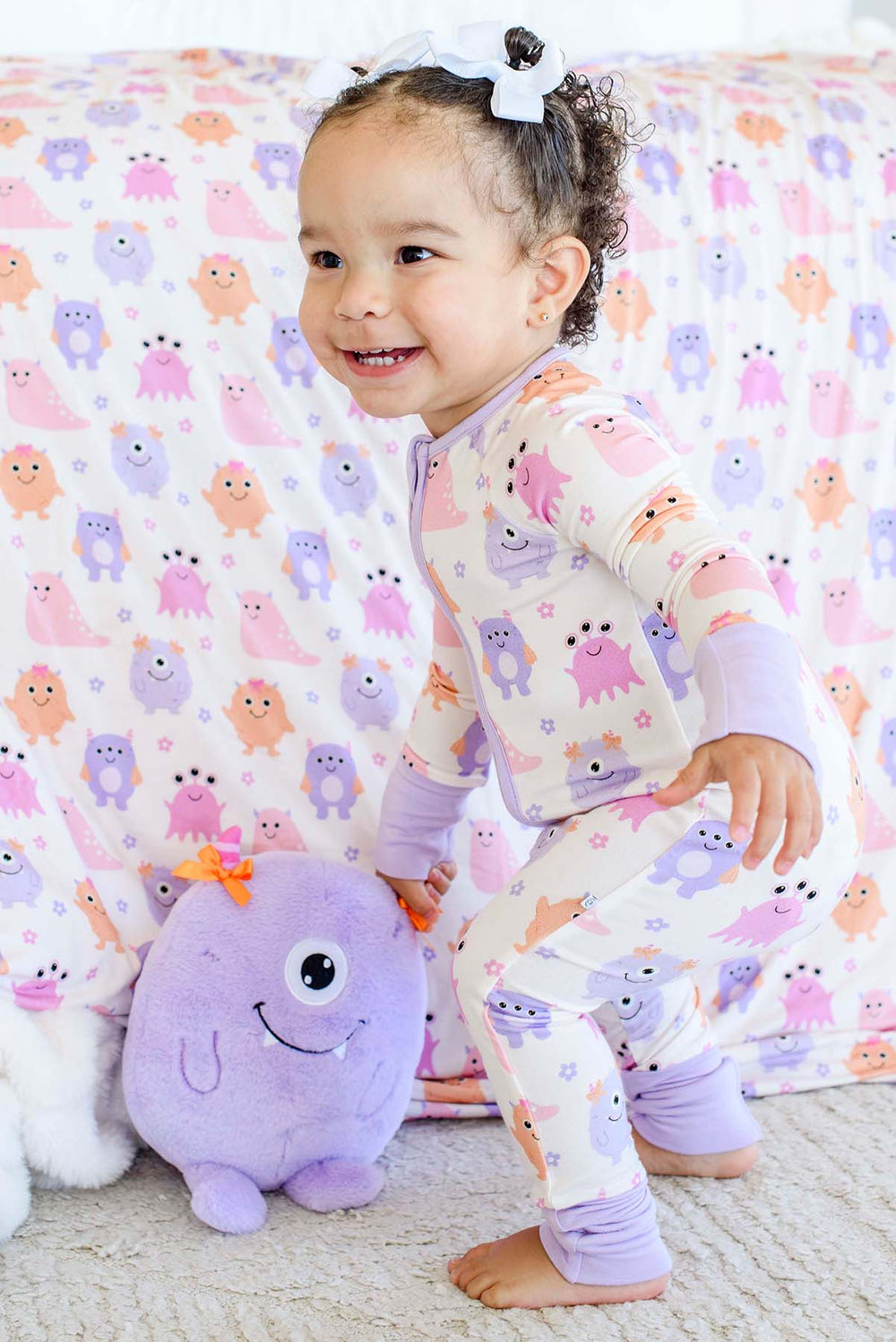 A toddler wearing a light purple and white long-sleeve pajama set with a colorful monster print, smiling and kneeling on a carpet next to a purple plush toy in front of a matching monster-print blanket.