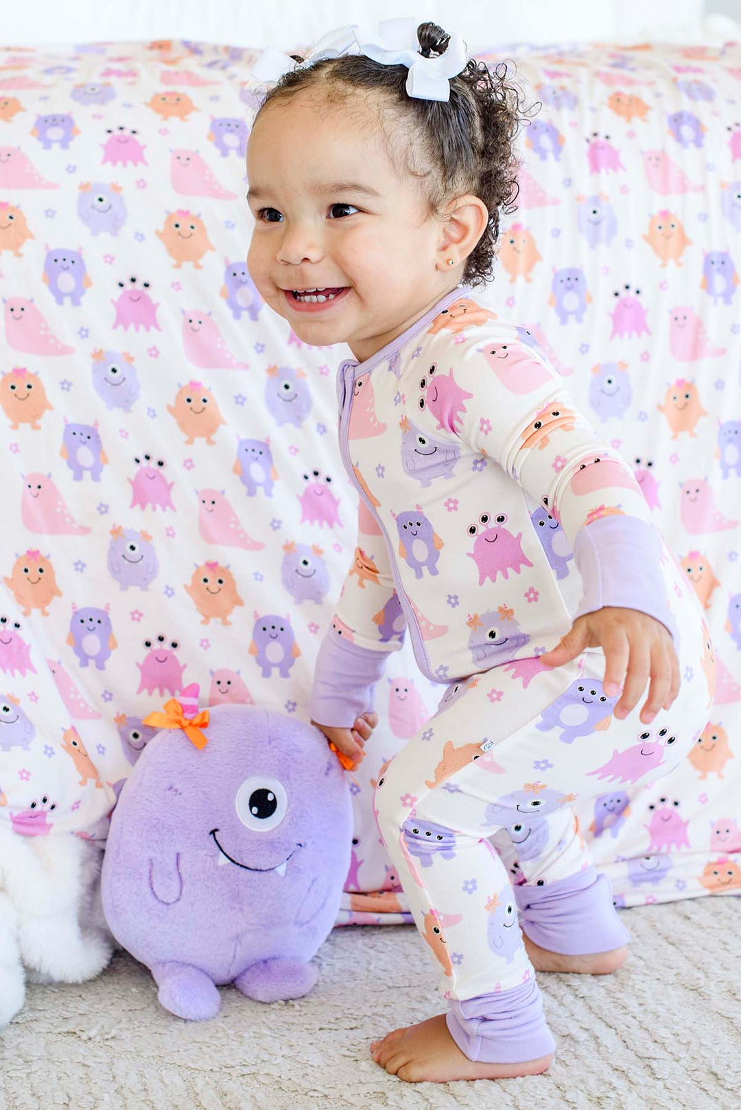 A toddler wearing a light purple and white long-sleeve pajama set with a colorful monster print, smiling and kneeling on a carpet next to a purple plush toy in front of a matching monster-print blanket.
