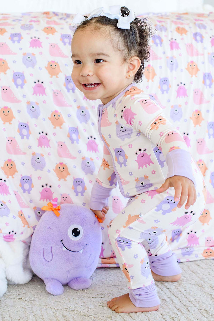 A toddler wearing a light purple and white long-sleeve pajama set with a colorful monster print, smiling and kneeling on a carpet next to a purple plush toy in front of a matching monster-print blanket.