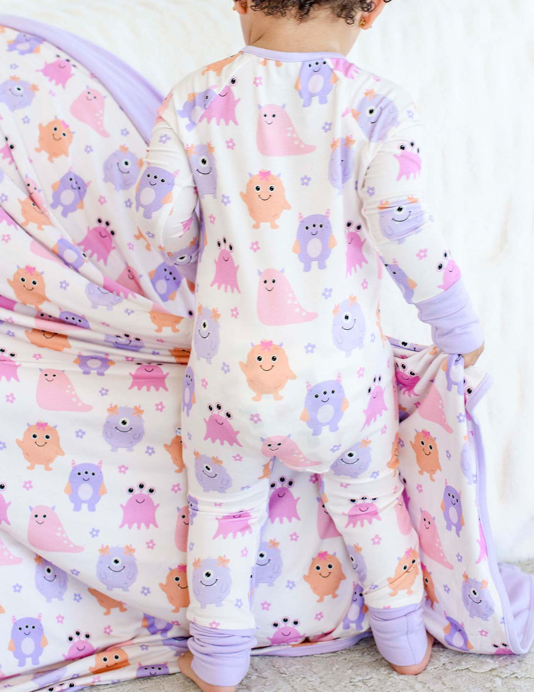 Back view of a toddler standing on a bed wearing a Caden Lane Little Monsters zipper footie, showing print detail and lavender trim against coordinating bedding