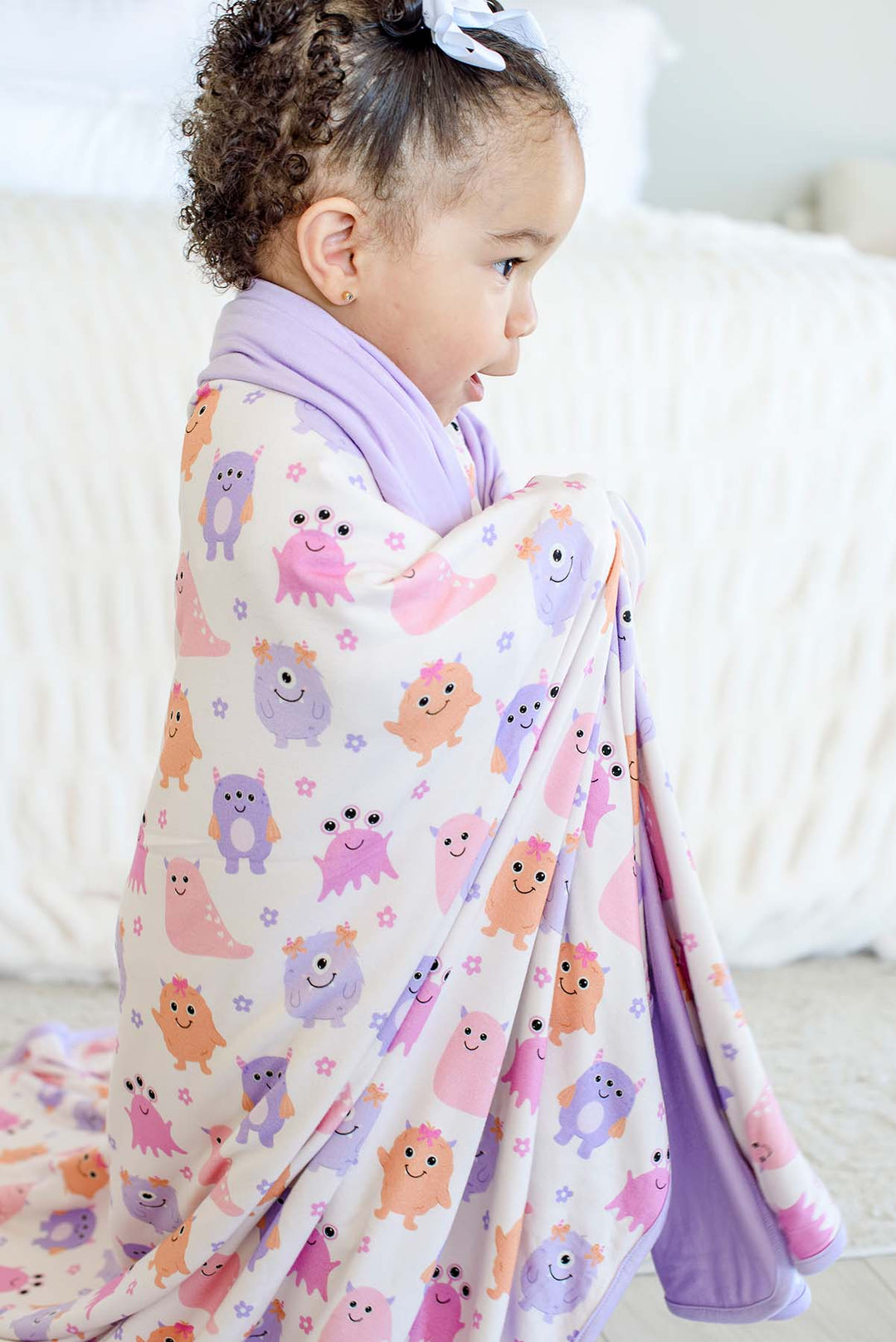 Toddler girl with curly hair and a white bow wrapped in a Caden Lane lavender-trimmed Little Monsters bamboo blanket, side profile