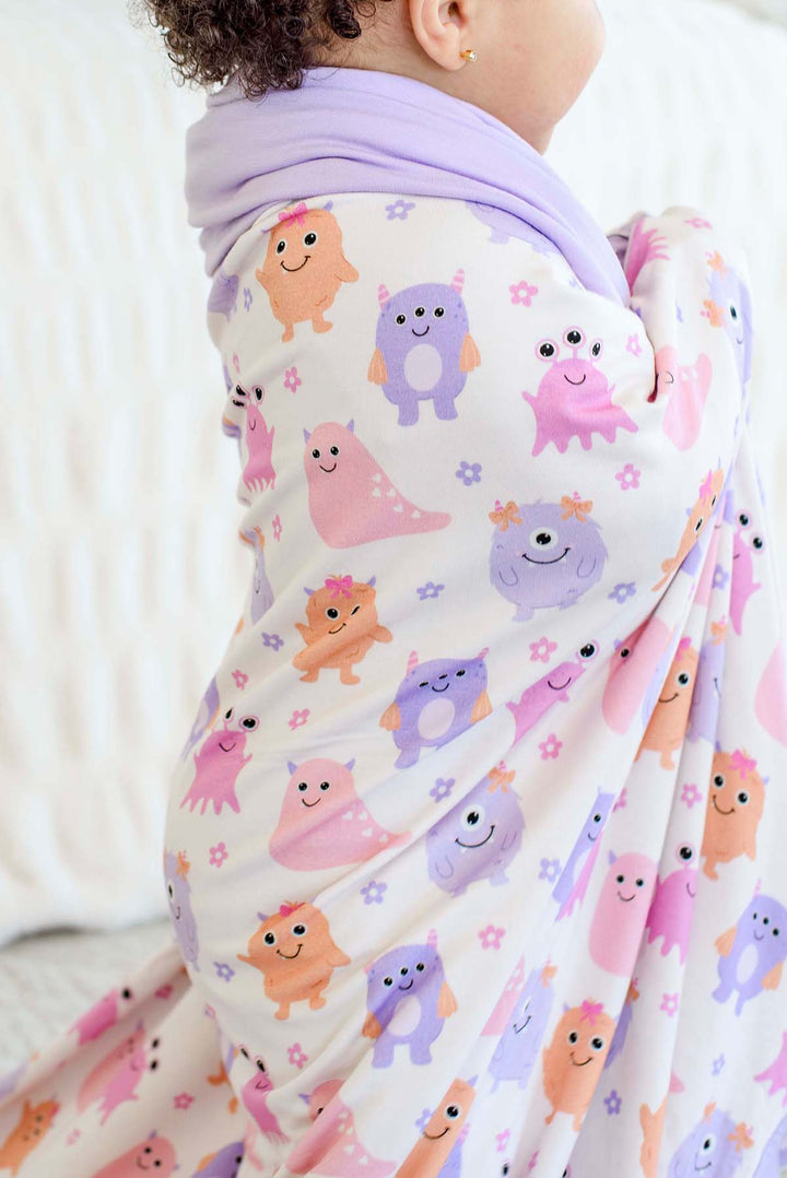 Close-up of a toddler wrapped in a Caden Lane lavender-trimmed Little Monsters bamboo blanket with pink, purple, and orange cartoon monsters